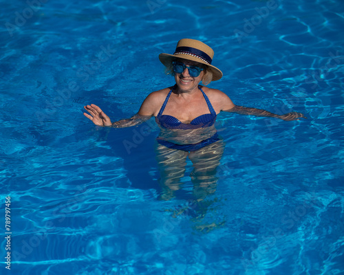 Portrait of an elderly woman in a hat and blue sunglasses swims in the pool. 