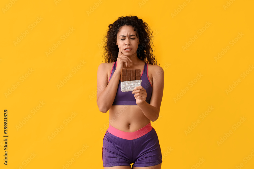 Thoughtful young African-American woman with sweet chocolate bar on yellow background - obrazy, fototapety, plakaty 