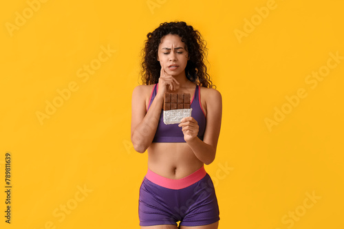 Thoughtful young African-American woman with sweet chocolate bar on yellow background