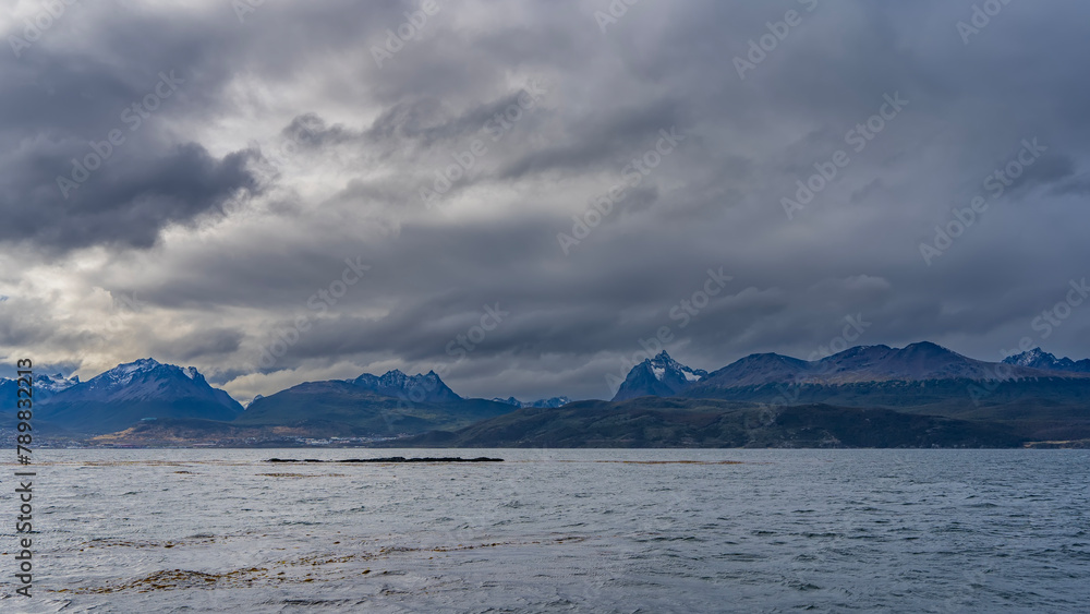 From the side of the Beagle Canal, the beautiful Andean Martial mountain range is visible. Snow-capped peaks against a cloudy sky. Ripples on the blue water of the strait. Argentina. 