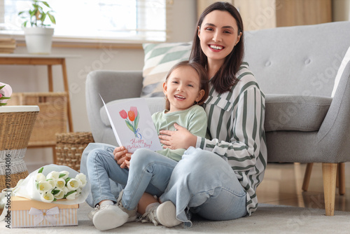 Happy mother with her cute little daughter and greeting card sitting on floor at home