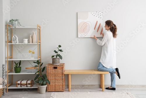 Young woman hanging picture on white wall in room