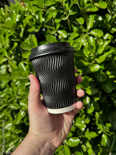 Hand holding black corrugated cardboard coffee cup with lid against background of lush green leafy bush. Urban eco friendly lifestyle. Blank space for sticker or label.