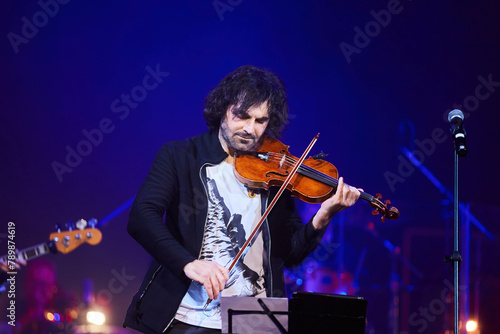 A violinist musician stands on stage with a violin in his hands during a concert. There is a bright light from the floodlights around.