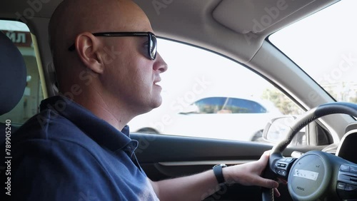 A close-up of a bald man in sunglasses is angry in the car, he honks while standing in a traffic jam. An angry man honks while sitting in a car, a traffic jam on the road.