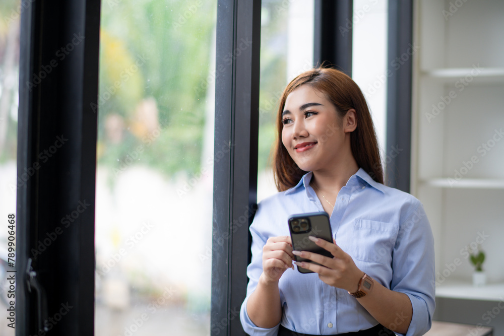 Smiling young Asian businesswoman using her smartphone by the window in a well-lit office, dressed in smart casual attire. Smiling young Asian businesswoman using her smartphone by the window in a well-lit office, dressed in smart casual attire.