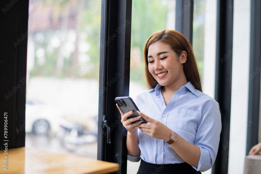 Smiling young Asian businesswoman using her smartphone by the window in a well-lit office, dressed in smart casual attire. Smiling young Asian businesswoman using her smartphone by the window in a well-lit office, dressed in smart casual attire.