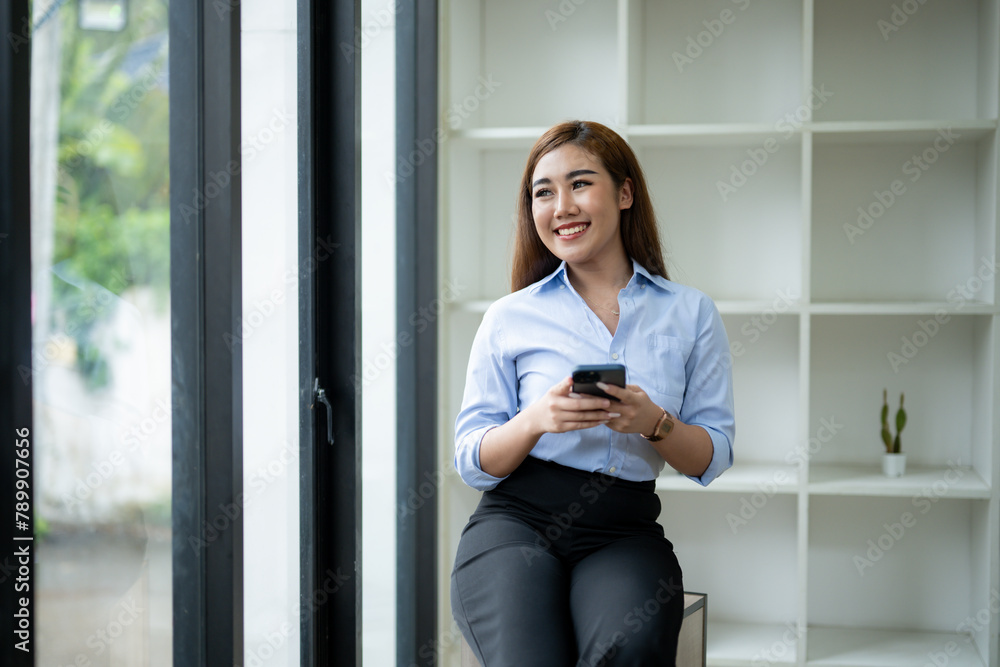 Smiling young Asian businesswoman using her smartphone by the window in a well-lit office, dressed in smart casual attire. Smiling young Asian businesswoman using her smartphone by the window in a well-lit office, dressed in smart casual attire.