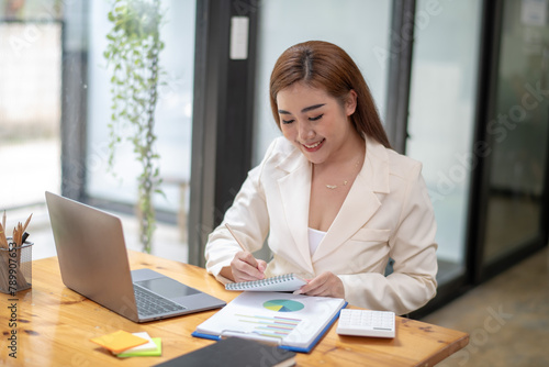 Professional Asian businesswoman typing on laptop at a wooden desk with paperwork and greenery in a bright office setting.