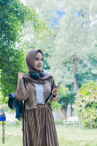 mature asian woman in veil candid smile carrying books and clipboard and bag standing in outdoor park. female students for the themes of education, technology and advertising