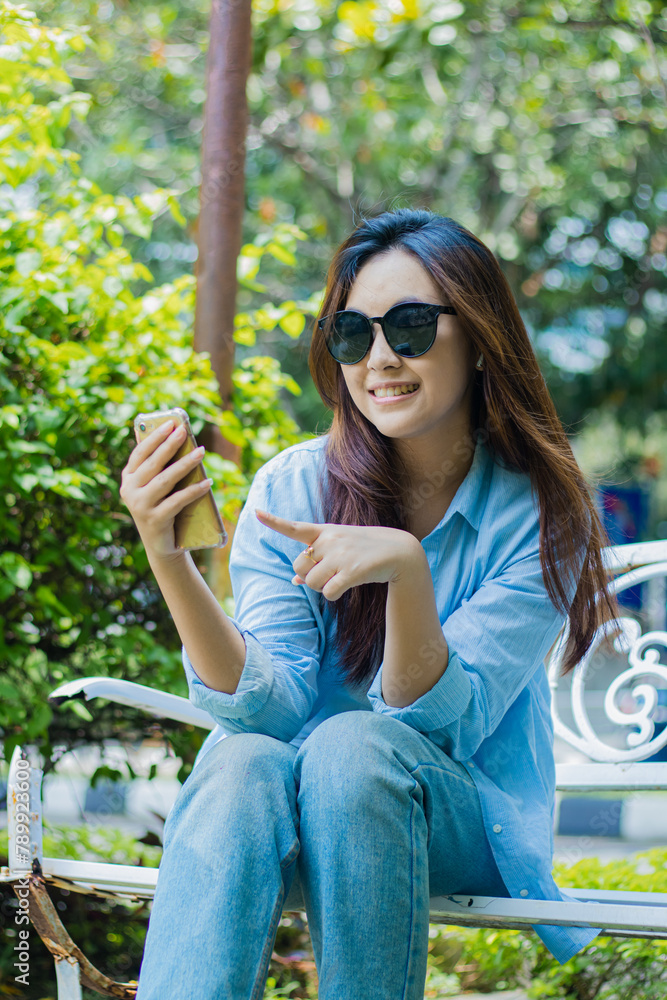 an Asian mature girl in sunglasses looks at the camera happily clicking discounts online shopping using a cell phone gadget sitting in an outdoor park. students for themes of technology and lifestyle an Asian mature girl in sunglasses looks at the camera happily clicking discounts online shopping using a cell phone gadget sitting in an outdoor park. students for themes of technology and lifestyle