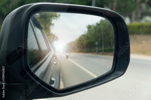 Inside view of mirrors wing. Rear view of a gray car with asphalt road and green trees in the daytime. Blurred image of a motorcycle approaching from behind.