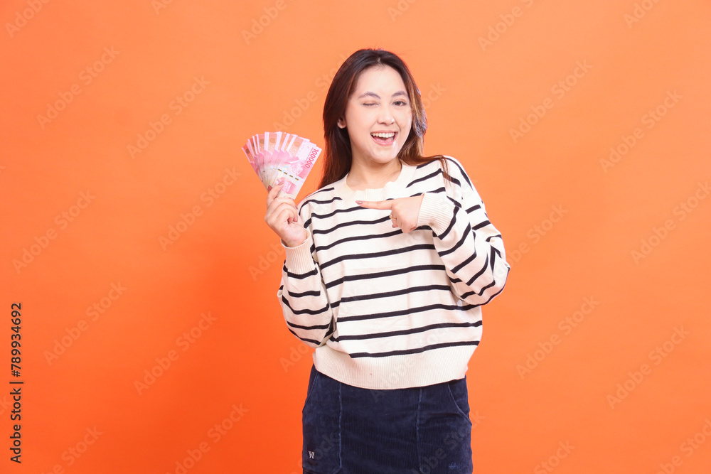 The expression of a cheerful Asian woman winks standing while holding some rupiah money for doing business. for fashion, business and finance concept The expression of a cheerful Asian woman winks standing while holding some rupiah money for doing business. for fashion, business and finance concept