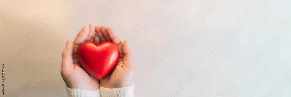 Obraz premium Cupped hands gently presenting a glossy red heart on a soft pastel background, symbolizing love and care.
