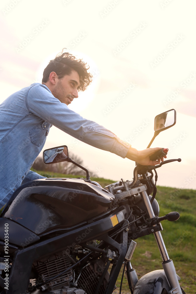 Curly haired Caucasian young man carrying his motorbike outdoors, looking away in direct sunlight, vertical image. Concept of lifestyle Curly haired Caucasian young man carrying his motorbike outdoors, looking away in direct sunlight, vertical image. Concept of lifestyle
