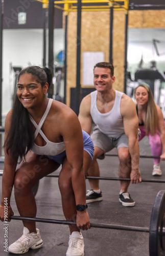 Group of multiracial smiling people doing dead lift with some bars with discs inside a gym, front view, sport dress, vertical image. Fitness and healthy lifestyle concept