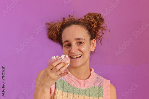 Pretty young caucasian woman smiling holding a donuts looking at camera and a candy colored T-shirt on pink background