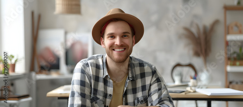 A jovial man is depicted in a studio setting. wearing a hat and sitting at his drawing board with a sketchpad visible on the right side of the frame.