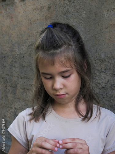 portrait of a nine year old girl standing near a stone wall and looking at her hands in the summer © Oleksandra
