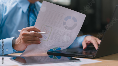 Businessman is sitting at a desk with a laptop and a piece of paper. He is holding a pen and looking at the paper. Concept of focus and concentration as the man works on a task