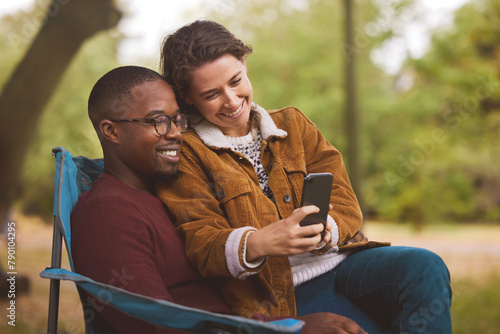 Camping, phone and smile with interracial couple in forest together for communication, hobby or leisure. Love, happy or relax with man and woman in nature for discovery, exploration or summer travel © peopleimages.com