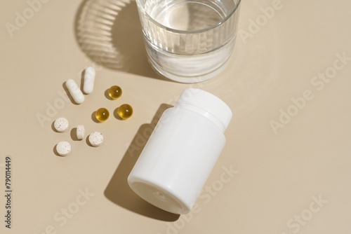 White mockup of blank jar and capsule pills on beige isolated background with glass of water. Health care, disease treatment, vitamins for beauty concept.