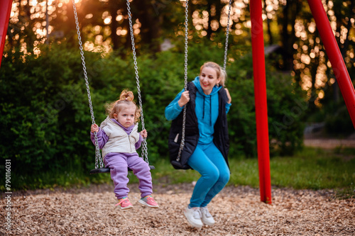A little girl and a woman enjoy swinging in a park at dusk, with golden sunlight filtering through trees in the background.