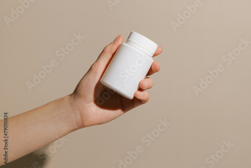 Female hand holding white small pill jar mockup on beige isolated background. Concept of pharmacy, health care, medicine, treatment. Image for your design.