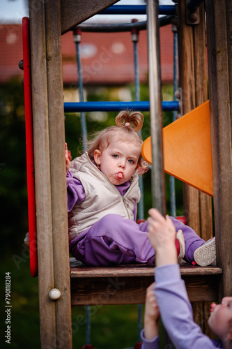 A playful child sticks out her tongue in a moment of fun, interacting with someone below her on a colorful playground tower