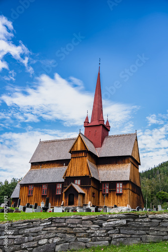 View of the 13th century medieval Ringebu Stave Church stands amidst headstones under a blue sky with clouds in Summer, echoing centuries of Norwegian heritage.