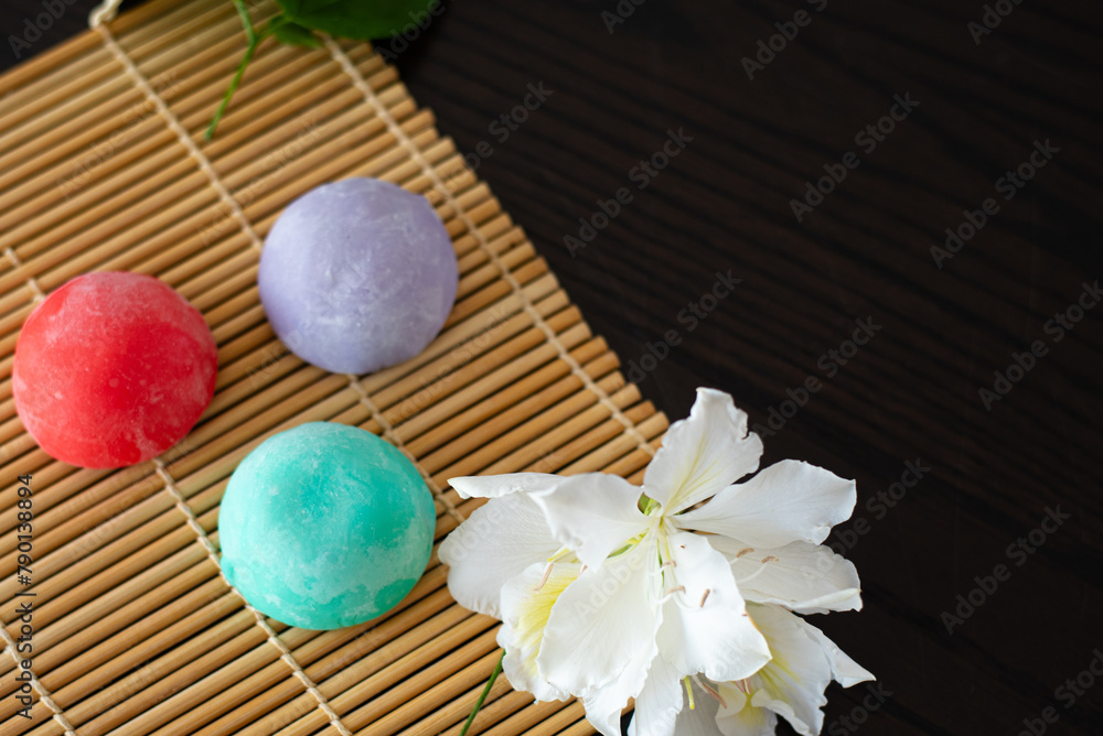 Three mochi different tastes and colors on bamboo makisu with flowers on black background. Japanese traditional frozen delicious dessert mochi. ice cream with dough of sticky rice. Asian cuisine. Three mochi different tastes and colors on bamboo makisu with flowers on black background. Japanese traditional frozen delicious dessert mochi. ice cream with dough of sticky rice. Asian cuisine.