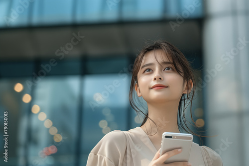 Hopeful Asian businesswoman with smartphone looking upward in city © wazamai
