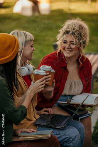 Three happy female students cheering with a coffee to go while sitting in a campus outdoors