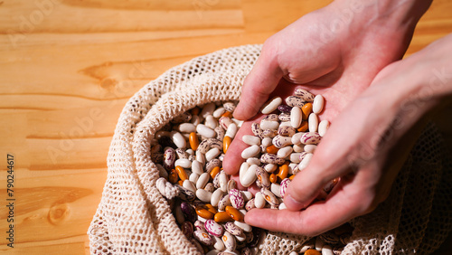 Hands pick through assortment of multicolored beans in crocheted bag on wooden table. Selecting quality produce. Nutritional choices. Conscious consumerism concept. Banner with copy space