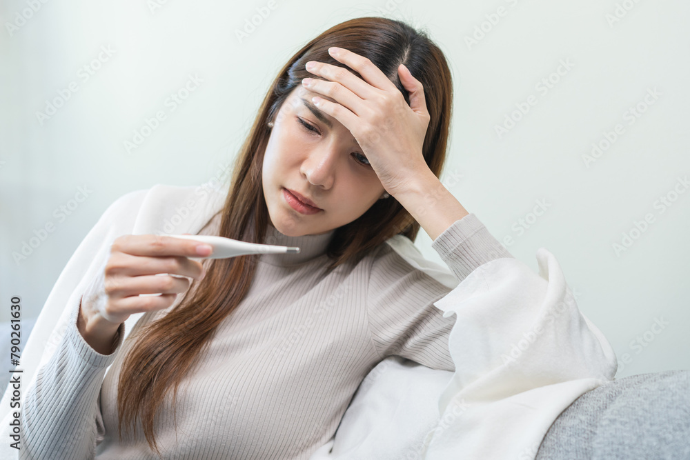 Sick, influenza asian young woman, girl headache have fever, hand touching forehead, holding thermometer for check measure body temperature, illness while sitting rest on sofa at home. Health care. Sick, influenza asian young woman, girl headache have fever, hand touching forehead, holding thermometer for check measure body temperature, illness while sitting rest on sofa at home. Health care.