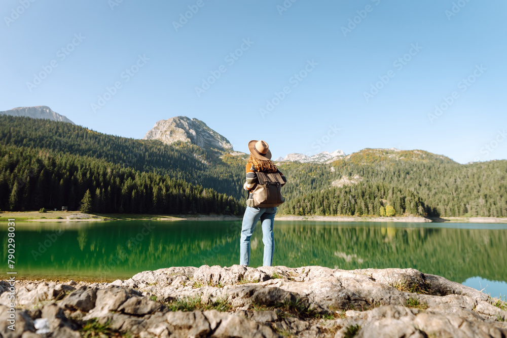 Obraz premium Back view young woman with backpack loking the view of the mountain lake in sunny weather. The concept of travel, vacation. Active lifestyle.