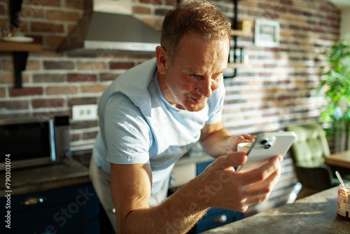 Middle aged man drinking coffee and checking smartphone in the kitchen photo