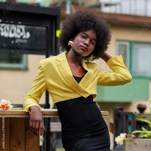 An attractive young African woman with a flower is posing outside in the city. She is wearing a black dress and a beautiful yellow jacket.