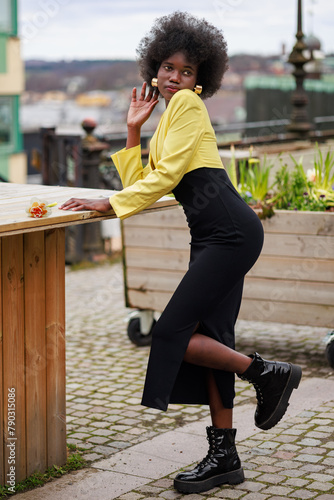 An attractive young African woman is posing outside in the city. She is wearing a black dress, a beautiful yellow jacket and boots.