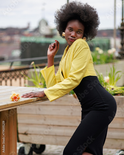 An attractive young African woman with a flower is posing outside in the city. She is wearing a black dress and a beautiful yellow jacket.