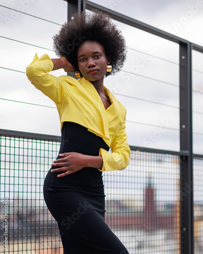 An attractive young African woman is posing on a footbridge with great view over the city. She is wearing a black dress and a beautiful yellow jacket.