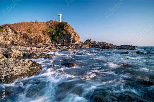 The sun sets behind a solitary lighthouse perched atop a cliff surrounded by a rugged, rocky coastline, evoking peaceful solitude. 