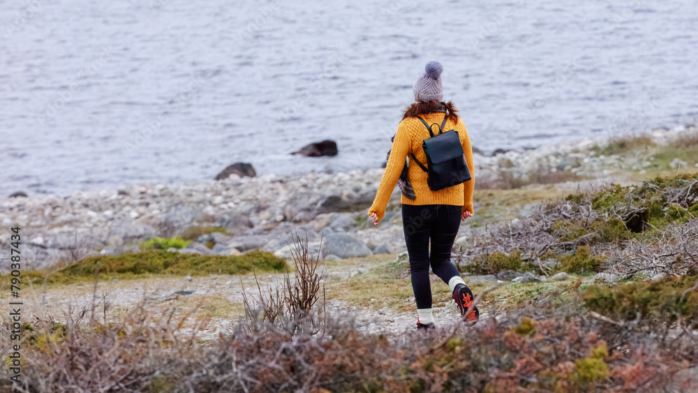 An active woman in a yellow sweater and winter accessories hiking by the sea, embodying the calm of a solitary coastal walk. Concept: meditation, solitude, sea exploration