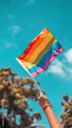 Vertical image of a colorful gay pride flag being waved on a blue sky background. LGBTQ