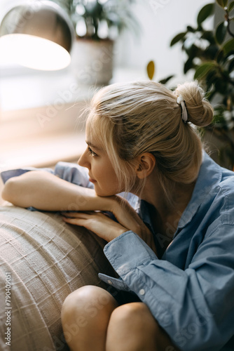 Young blonde woman sitting on sofa at home, looking through the window.