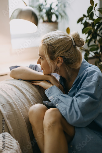 Young blonde woman with a hair bun, sitting on sofa at home, looking through the window.