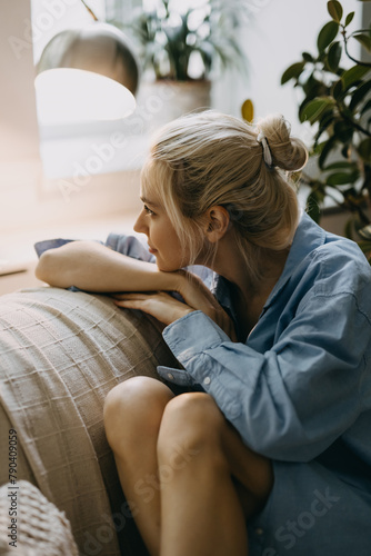 Obraz na plátně Young blonde woman with a hair bun, sitting on sofa at home, looking through the window