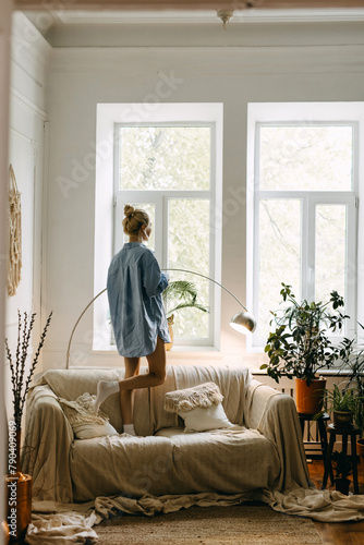 Woman in blue oversized shirt standing on sofa in a sunlit living room, looking at the window.