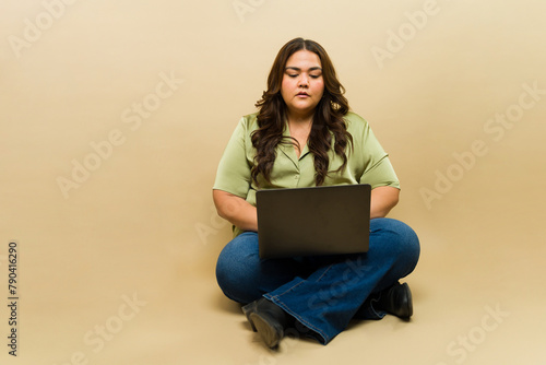 Thoughtful plus-size woman using a laptop on a beige background in a studio setting