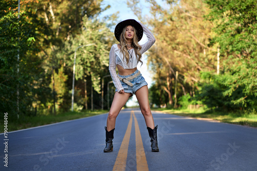 Portrait of a stand woman in a cowboy hat, white shirt, jeans short pants and black boots, on a paved road with trees at outdoors in a sunny afternoon.